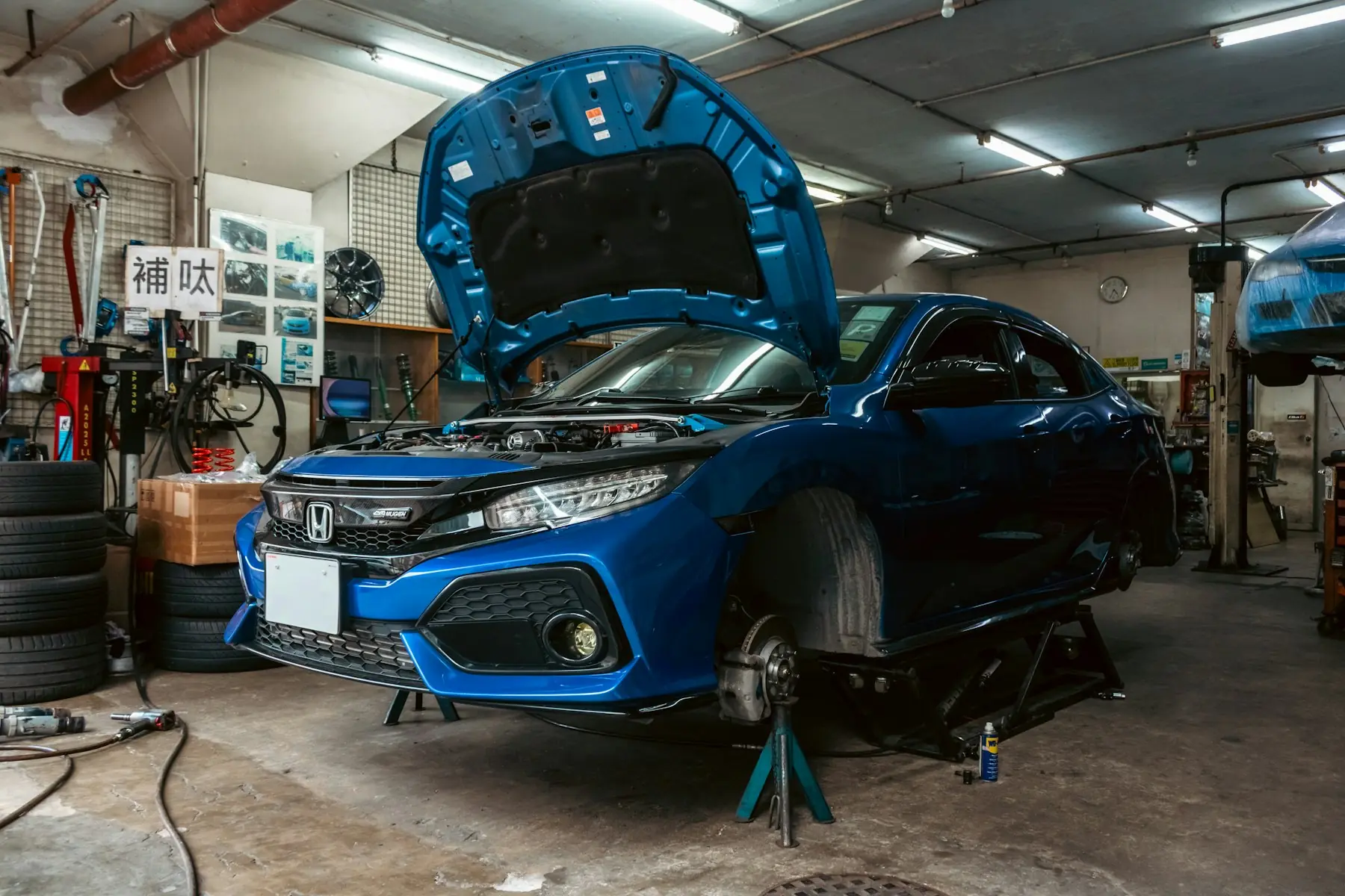 A blue sedan on jack stands with its hood up in a working repair bay