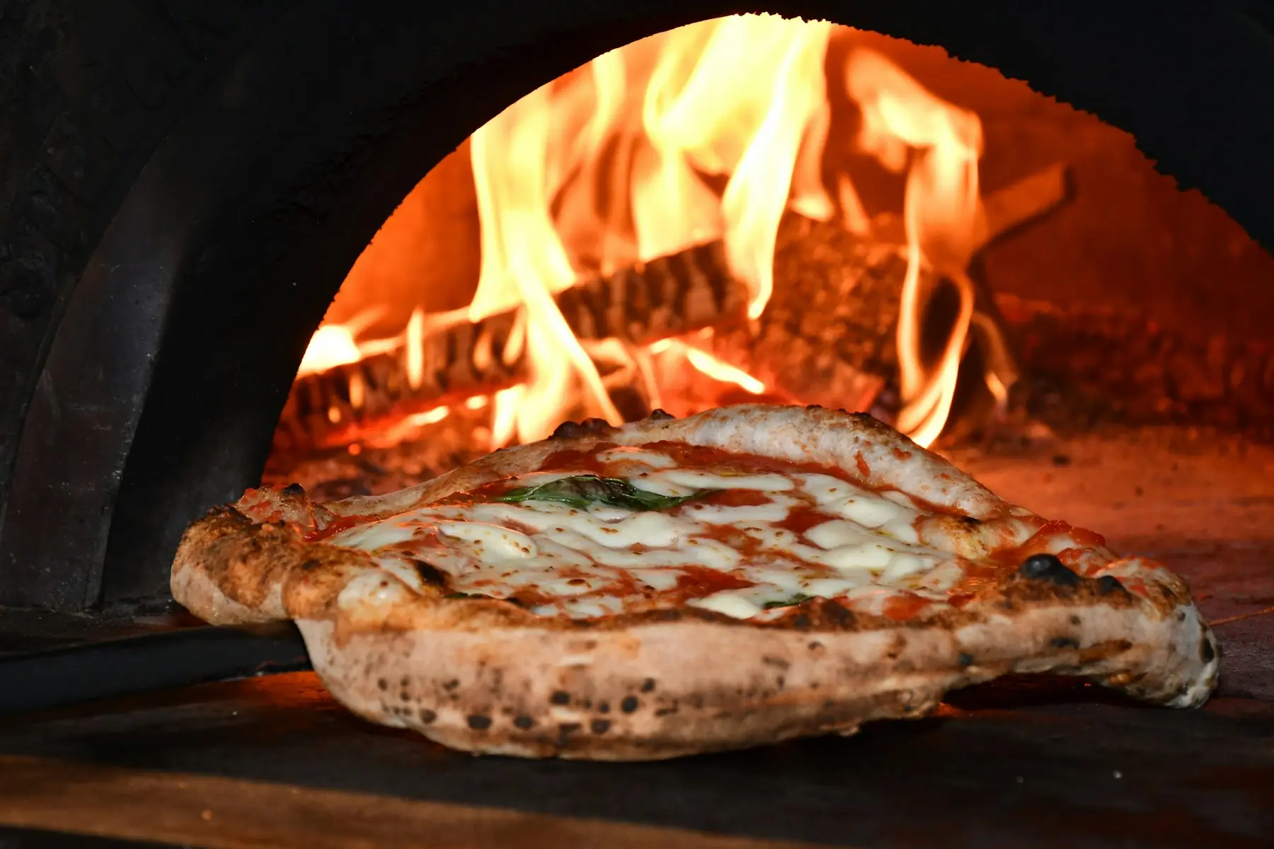 A margherita pizza blistering beside the flames in a wood-fired oven